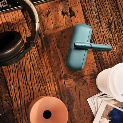 Türkises IQOS-Gerät liegt auf einem Holztisch, umgeben von Kopfhörern, einem Coffee-to-go-Becher und verstreuten Fotos.
