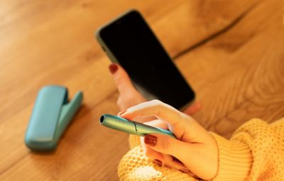 Person hält IQOS ORIGINALS DUO Turquoise in der einen und Smartphone in der anderen Hand.