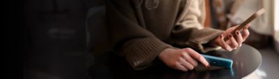 Frau in braunem Pullover hält IQOS ORIGINALS ONE in Turquoise und Smartphone vor dunklem Hintergrund.