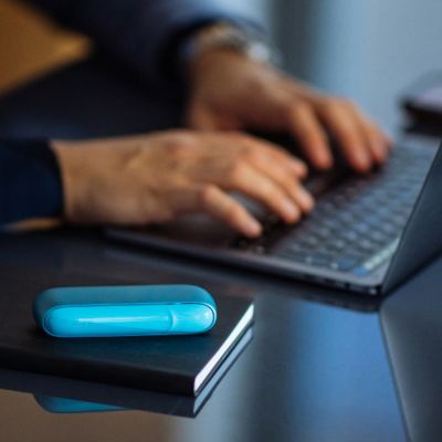 IQOS ORIGINALS DUO Turquoise auf schwarzem Notizbuch, im Hintergrund tippt eine Person am Laptop.
