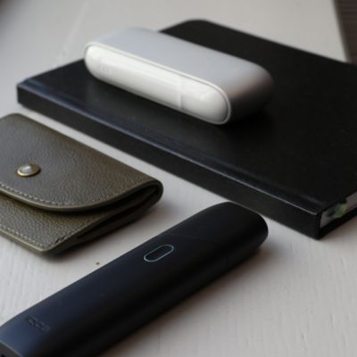 IQOS ORIGINALS DUO Silver auf schwarzem Notizbuch. Davor graue Geldbörse und IQOS ORIGINALS ONE in Slate.