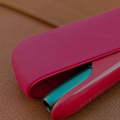 Étui rouge texturé ouvert, laissant apparaître un dispositif électronique IQOS de couleur turquoise, posé sur une surface marron.