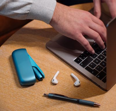 Ein blaues IQOS ORIGINALS DUO liegt auf einem Tisch mit einem Laptop, Kopfhörern und einem Stift.