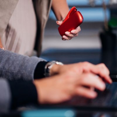 Person tippt im Vordergrund am Laptop, im Hintergrund hält weitere Person ein IQOS ORIGINALS DUO Scarlet in ausgestreckter Hand.