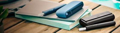 Two iqos ILUMA Devices resting on a wooden table, one of which rests on a blue notebook