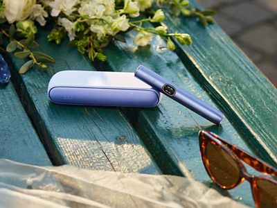 Ein IQOS ILUMA i in Violet liegt neben einem Blumenstrauß und einer Sonnenbrille auf einer Parkbank.