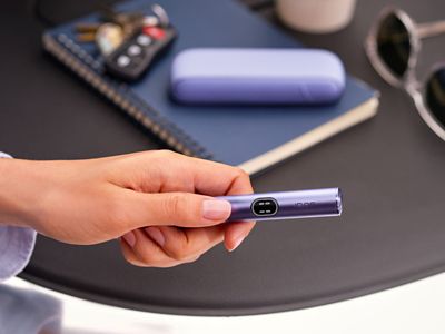 Ein IQOS ILUMA i Gerät in der FarbeViolet, im Hintergrund ein Notizbuch, eine Sonnenbrille und Autoschlüssel. 
