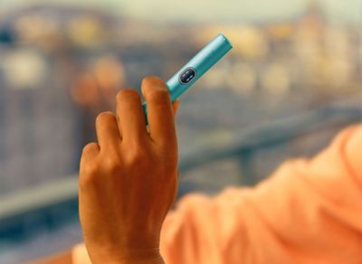 Hand hält ein türkises IQOS ILUMA i Gerät mit Touchscreen im Freien.
