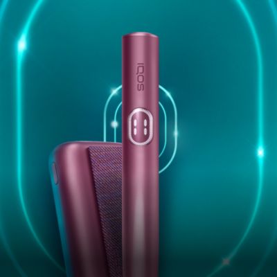Zařízení IQOS ILUMA i PRIME v barvě Garnet Red a jeho nabíječka, na tmavě tyrkysovém pozadí