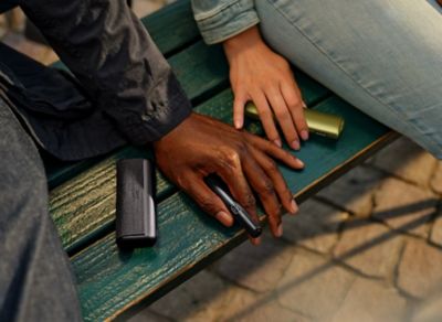 IQOS ILUMA i PRIME in der Farbe Midnight Black und IQOS ILUMA i ONE in Leaf Green liegen auf Parkbank neben Händen