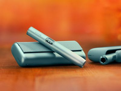 Držalo IQOS ILUMA i PRIME v svetlomodri barvi Breeze Blue na žepnem polnilniku, z napravo IQOS ILUMA i v ozadju.