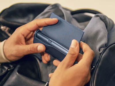 Nahaufnahme zweier Hände die ein geöffnetes IQOS ILUMA i PRIME in der Hand halten, im Hintergrund eine Reisetasche.