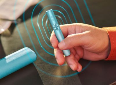 Ruka držící modré zařízení IQOS ILUMA i PRIME; nabíječka leží na stole