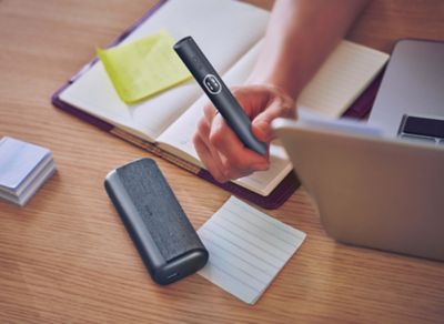 Hand hebt IQOS ILUMA i PRIME Midnight Black Holder von Schreibtisch auf