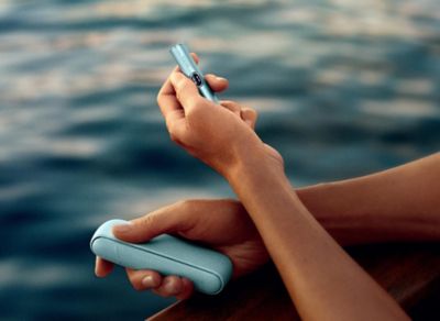 IQOS ILUMA i in der Farbe Breeze Blue in Händen am Wasser.