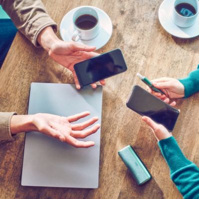 Dos personas con teléfonos móviles sentadas a la mesa; una de ellas también sostiene un dispositivo IQOS ILUMA PRIME color jade blue. 