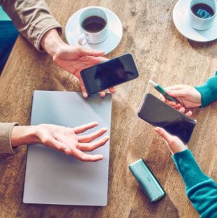 Two people holding mobile phones sit at a table; one also holds a jade blue IQOS ILUMA PRIME device.