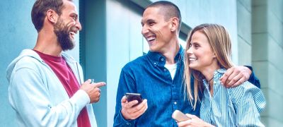 Two people holding IQOS ILUMA devices smile as their friend looks at the IQOS app on her mobile phone.