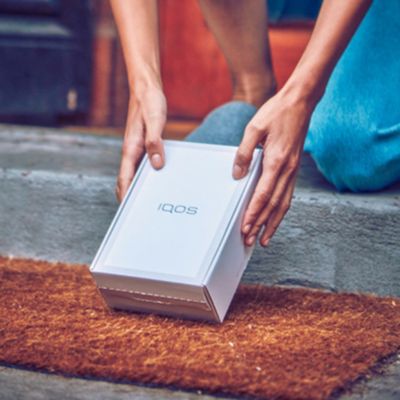 Two hands lifting an IQOS box from a front door mat.