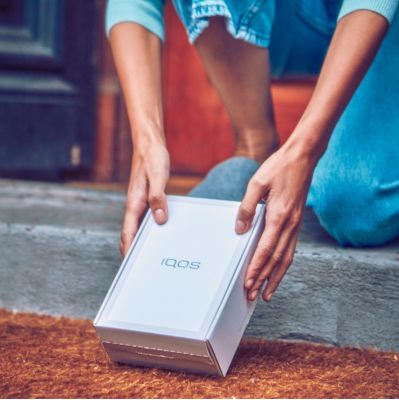 Two hands lifting an IQOS box from a front door mat.
