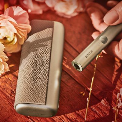 Dispositivo IQOS ILUMA PRIME color Gold Khaki colocado en un puesto de flores.