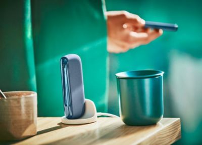 Un hombre sostiene en sus manos un IQOS ILUMA azul, con Holder y una taza en la mesa