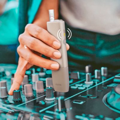A DJ adjusts dials on a turntable while holding an IQOS ILUMA ONE.