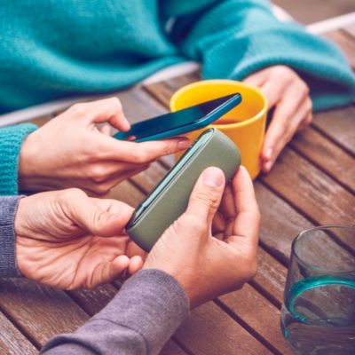 Hände halten ILUMA MID Moss Green Tabakerhitzer neben Smartphone