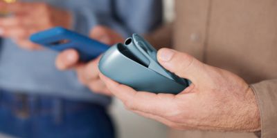 Ein Mann hält ein geöffnetes IQOS Originals Duo Charger und ein Smartphone in der anderen Hand.