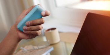 Frauenhände die ein IQOS Originals Duo an einem Arbeitstisch halten.
