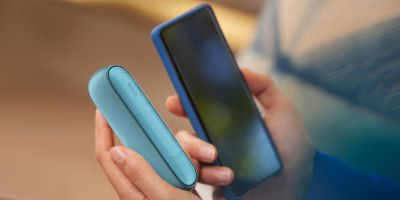 Menschenhände die ein IQOS Originals Duo halten und gleichzeitig ein Smartphone bedienen.