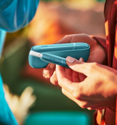 Hands holding IQOS Originals Duo heated tobacco holder and pocket charger in turquoise color.