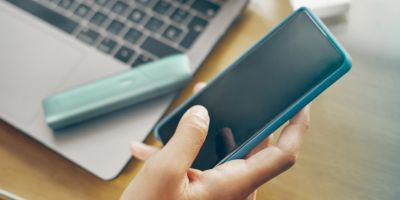 Frauenhänd die ein Smartphone über einem Laptop und einem IQOS Originals One bedienen.