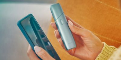 Frauenhände die ein IQOS Originals One und ein Smartphone bedienen.