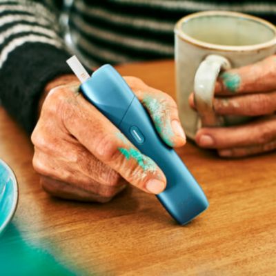 Hands with paint holding a coffee cup and a turquoise IQOS Originals One heated tobacco device.