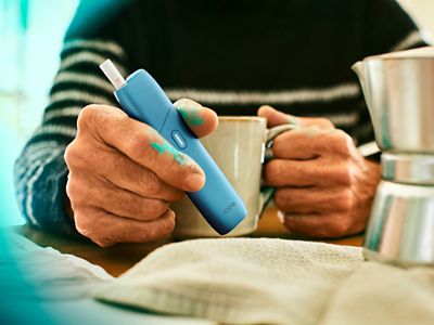 Hands holding a coffee cup and a turquoise IQOS ORIGINALS ONE device