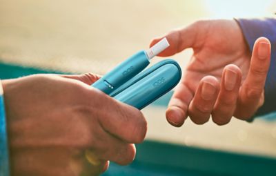 Hands exchanging a turquoise IQOS Originals Duo heated tobacco device.