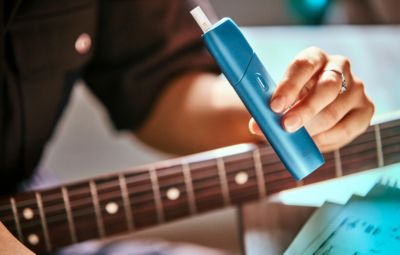 Hand holding turquoise IQOS Originals ONE heated tobacco device with a guitar behind.