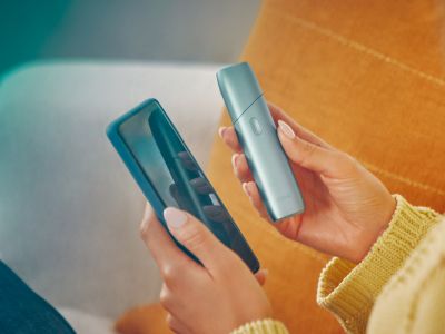 Femme tenant dans une main l'appareil IQOS Originals One en turquoise et dans l'autre un téléphone portable.