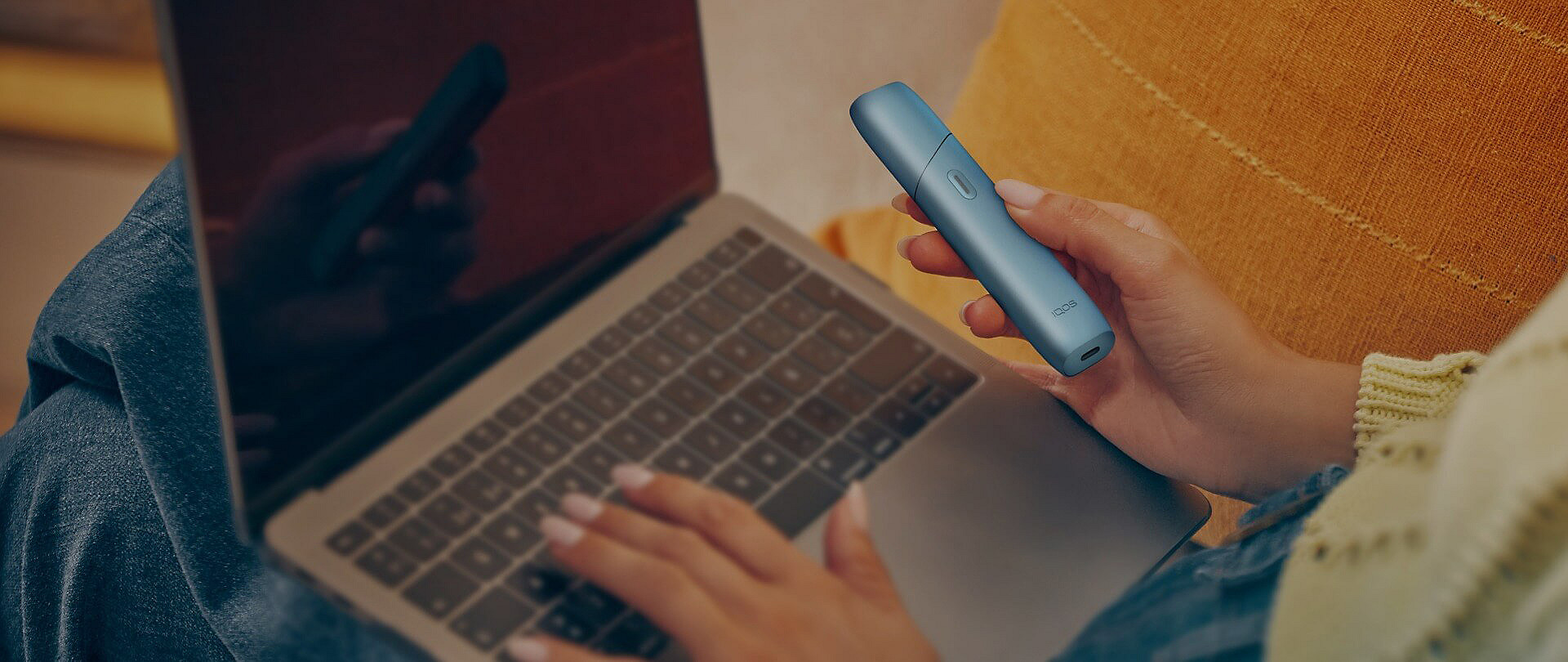 Woman holding an IQOS Originals ONE heated tobacco device and typing on laptop