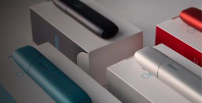 Mehrere IQOS ORIGINALS ONE Geräte in verschiedenen Farben liegen nebeneinander auf einer Verpackung.