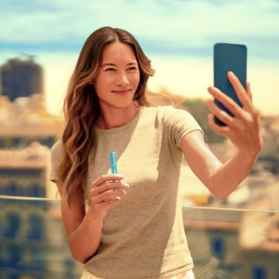 Woman with long wavy hair taking a selfie outdoors, showcasing an ILUMA i holder with a blurred urban skyline in the background.