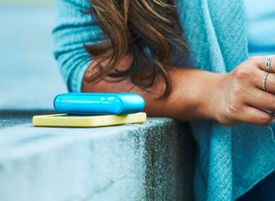 Person sitzt auf Betonstufe, vor ihr liegt ein Smartphone in gelber Hülle und ein IQOS ORIGINALS DUO Turquoise.