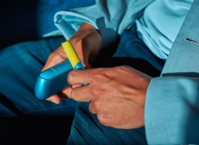 Mann im blauen Anzug hält IQOS ORIGINALS DUO Turquoise mit gelber Kappe in den Händen.