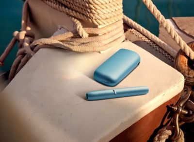 Türkiser IQOS-Charger und Holder liegen auf einem Bootselement neben Seilen und Wasser.