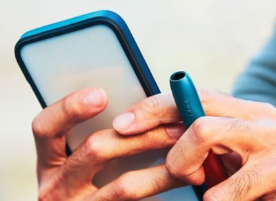 Person hält ein Smartphone mit Hülle und einen blauen IQOS ORIGINALS DUO Pocket-Charger in der Hand.