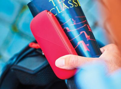 Hand hält IQOS ORIGINALS DUO Scarlet mit roter Silikonhülle und ein Magazin vor unscharfem Hintergrund.
