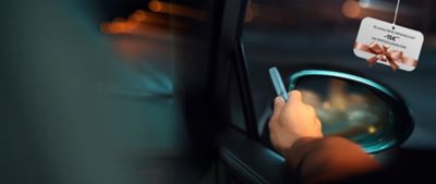 Eine Hand hält ein IQOS Gerät nahe einem Autofenster bei Nacht, darüber hängt ein Geschenkanhänger.