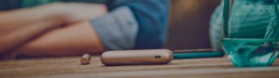 A close-up of an IQOS 3 DUO device resting on a table, in the background a person sits with their arms folded