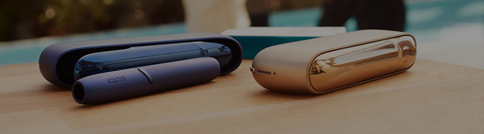 Two IQOS 3 Devices resting on a wooden table, in the blurred background we can see a group of people
